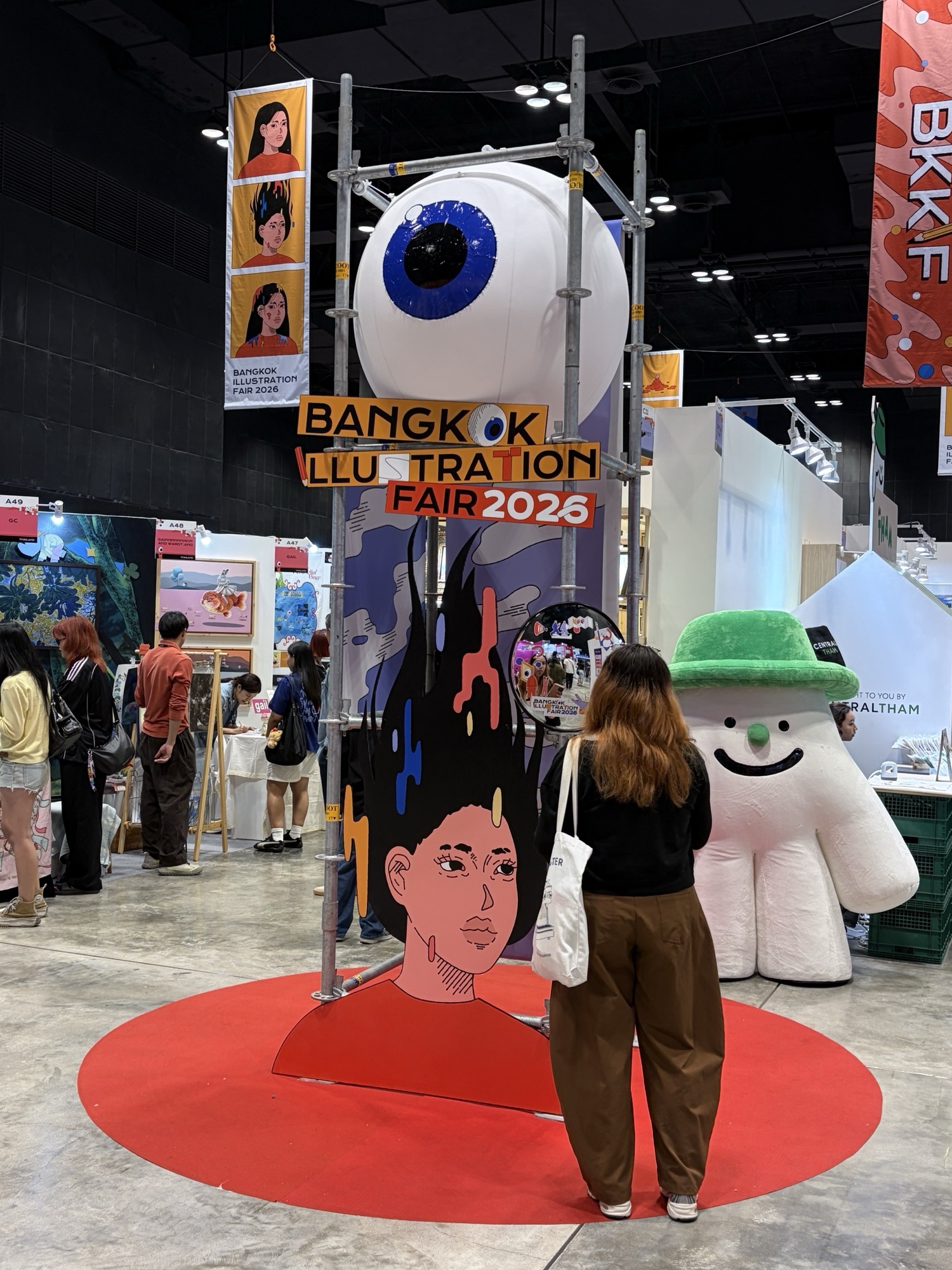 ป้าย Bangkok Illustration Fair 2026 บนนั่งร้าน มีลูกตาเป่าลม และป้ายหน้าผู้หญิงผมดำมีสีหยด