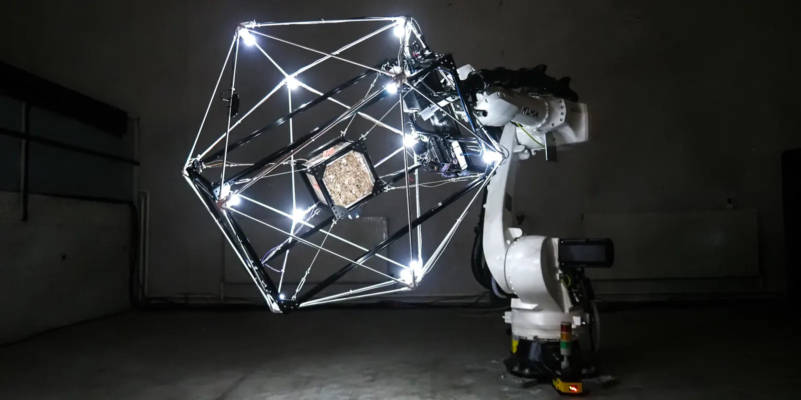 ภาพรวมของงาน MycoGravity โครงไฟทรงหลายเหลี่ยมติดอยู่บนแขนหุ่นยนต์ KUKA ในห้องมืด