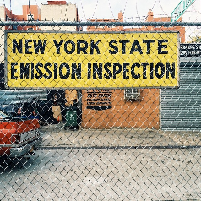 ป้ายเหลืองเขียนว่า NEW YORK STATE EMISSION INSPECTION หลังรั้วลวดตาข่าย