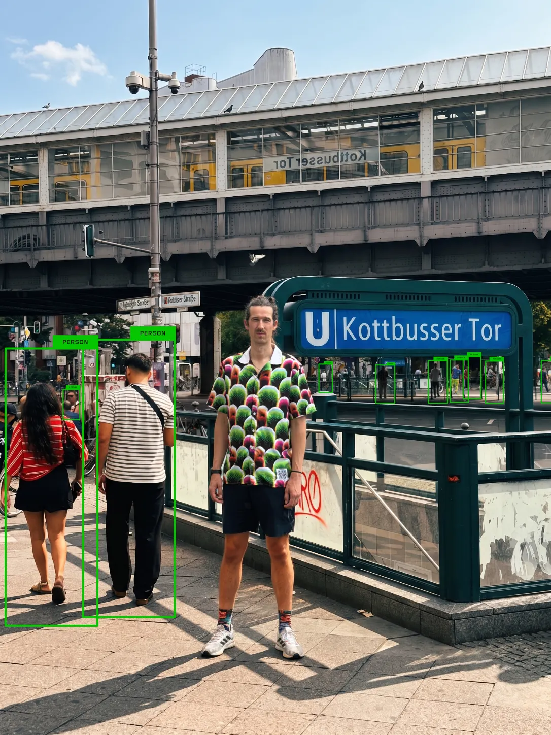 ชายใส่เสื้อ Digital Camouflage ยืนหน้าป้ายสถานี Kottbusser Tor ในเบอร์ลิน คนรอบข้างมีกรอบตรวจจับขึ้น
