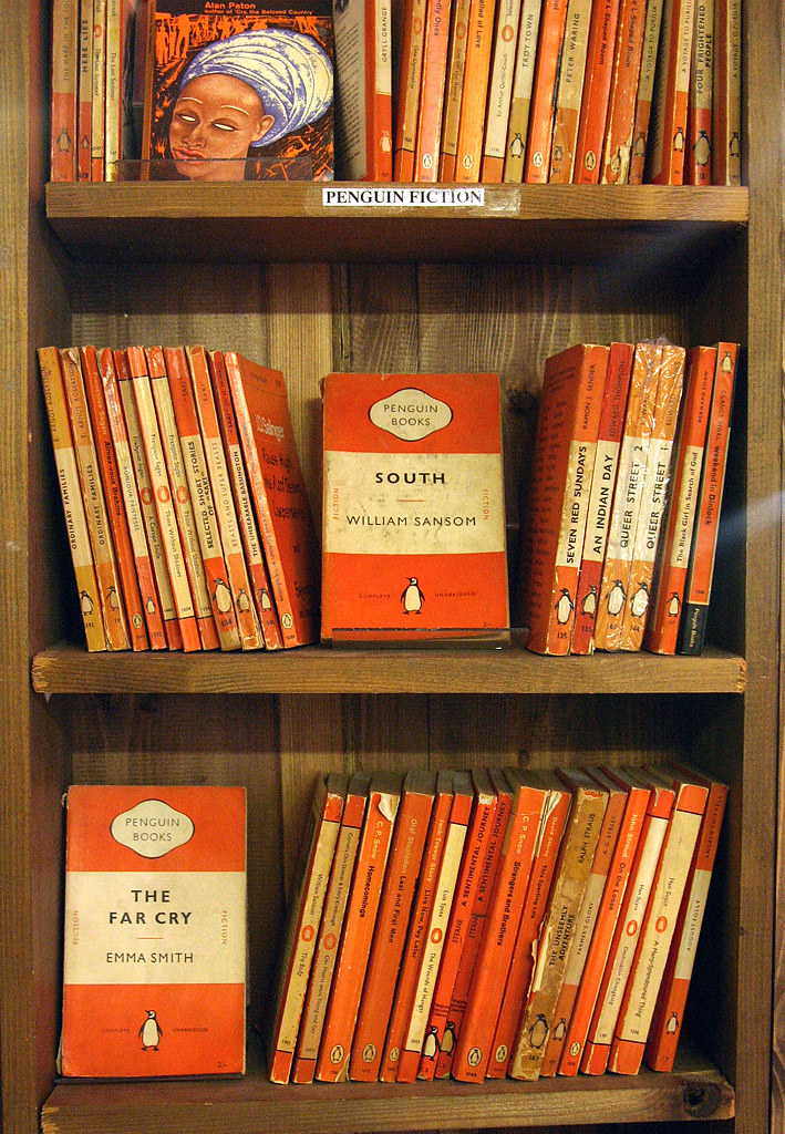 ชั้นหนังสือปกอ่อน Penguin รุ่นเก่าสีส้ม เรียงกันหลายสิบเล่ม ปกใช้แถบสามชั้นและตัวอักษร Gill Sans