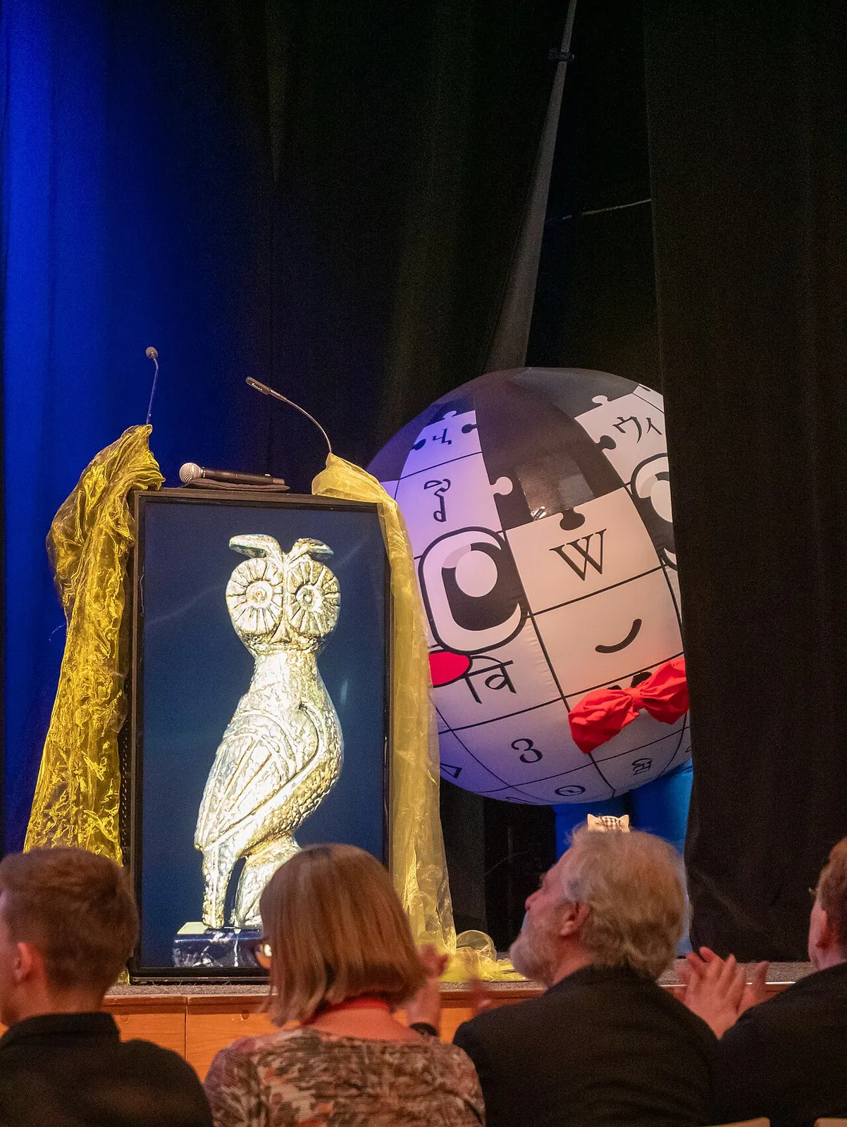 Baby Globe ในร่างลูกโป่งยักษ์ยืนบนเวที WikiCon 2026 ที่เมืองเรเกนส์บวร์ค ข้างรูปนกฮูกทอง