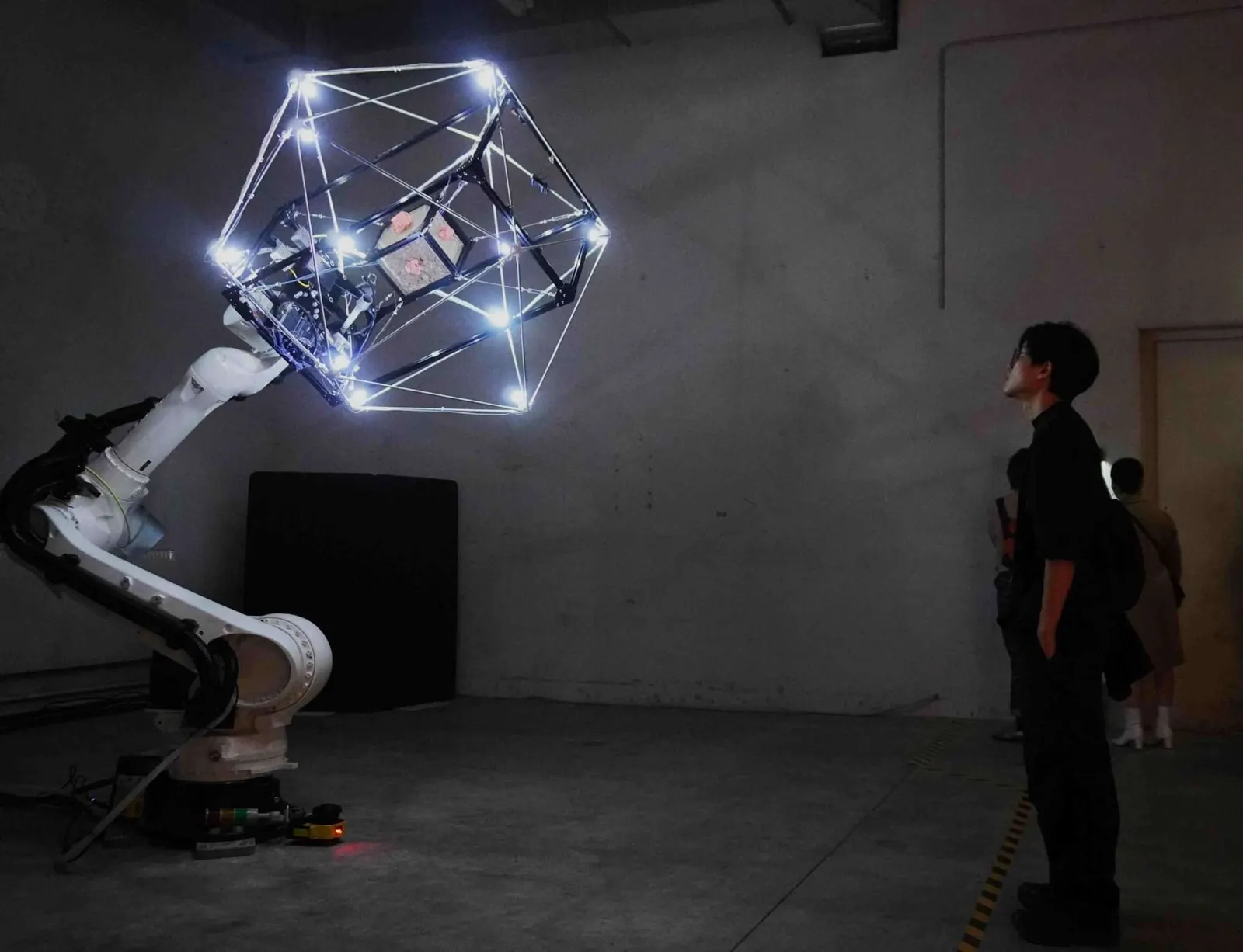 คนดูยืนมองโครงไฟของ MycoGravity ที่ลอยอยู่บนแขนหุ่นยนต์ในห้องนิทรรศการ