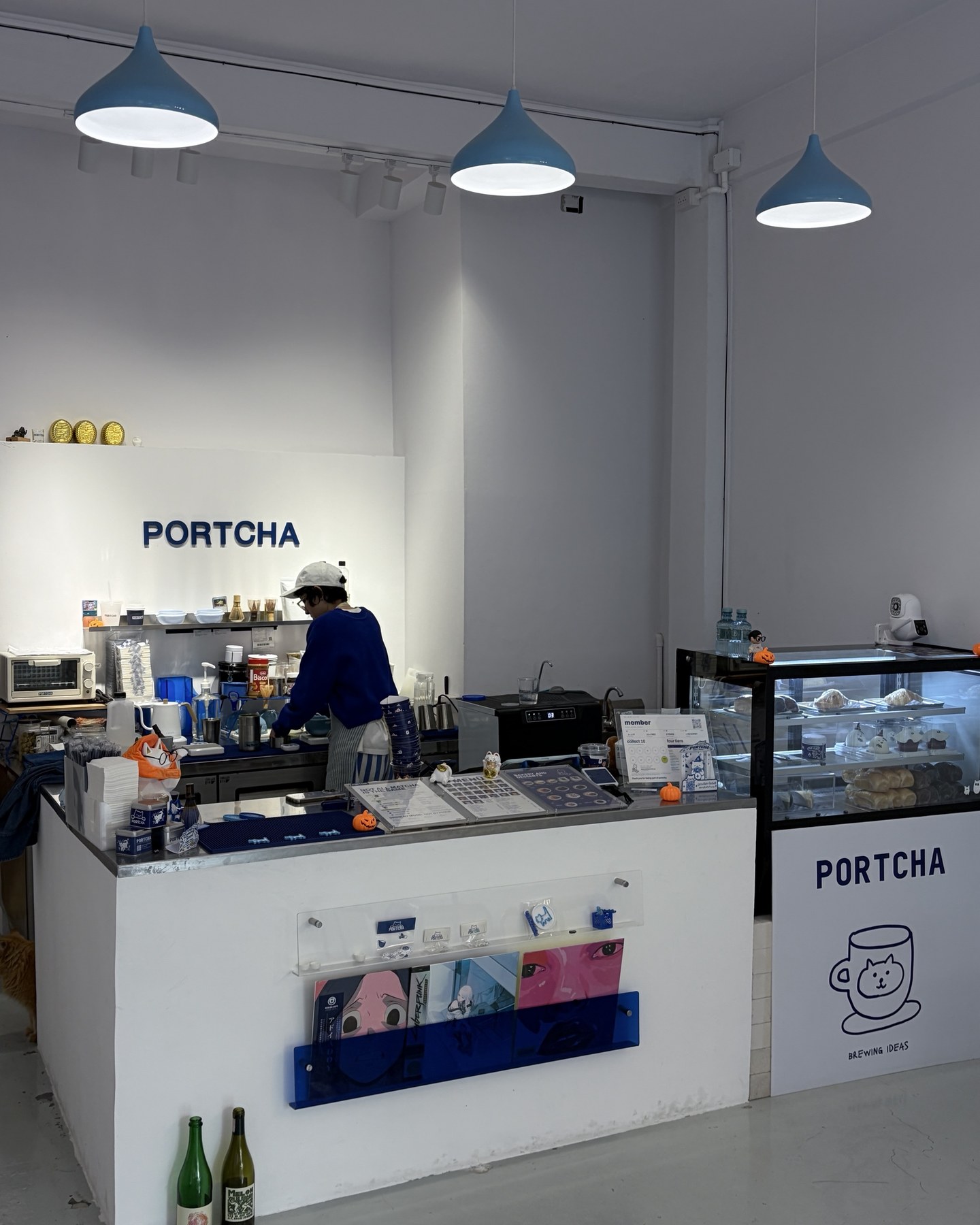เคาน์เตอร์ของ Portcha Cafe ป้าย PORTCHA สีน้ำเงินบนผนังขาว กับโคมไฟสีฟ้าสามดวง