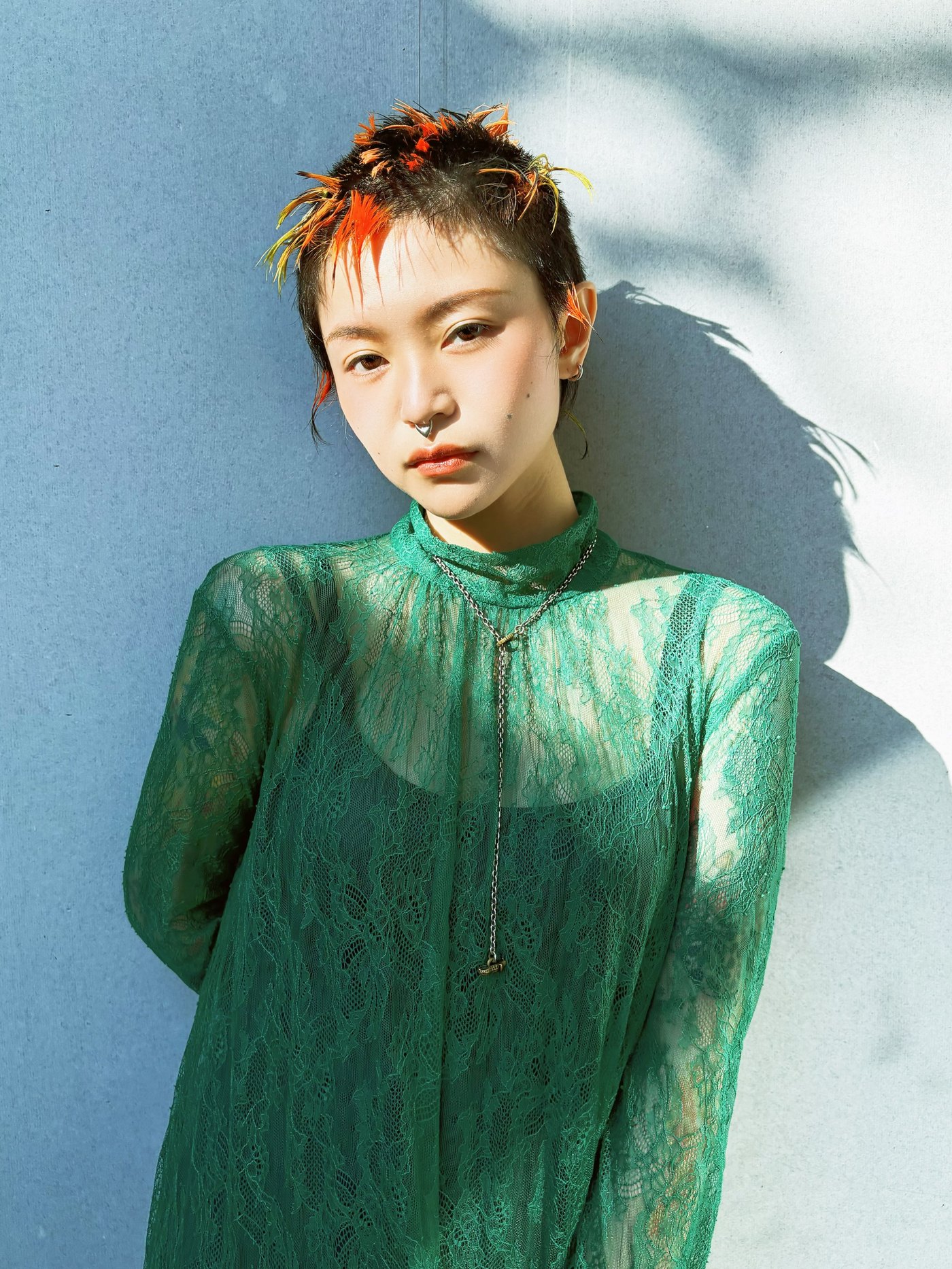 นางแบบผมสั้นสีน้ำตาลแซมแดงส้ม ใส่เสื้อลูกไม้สีเขียว ยืนพิงผนังในแสงแดด