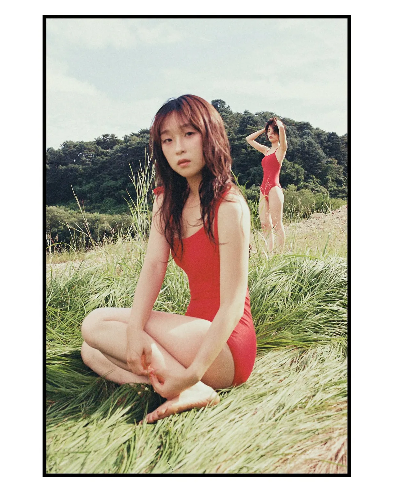 ผู้หญิงในชุดว่ายน้ำสีแดงนั่งขัดสมาธิบนหญ้า มีอีกคนยืนอยู่ด้านหลังไกลออกไป ภาพใส่กรอบดำหนา