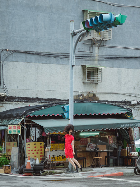 หญิงชุดแดงลอยตัวที่มุมถนนกับเสาไฟจราจรและแผงตลาด โดย Natsumi Hayashi